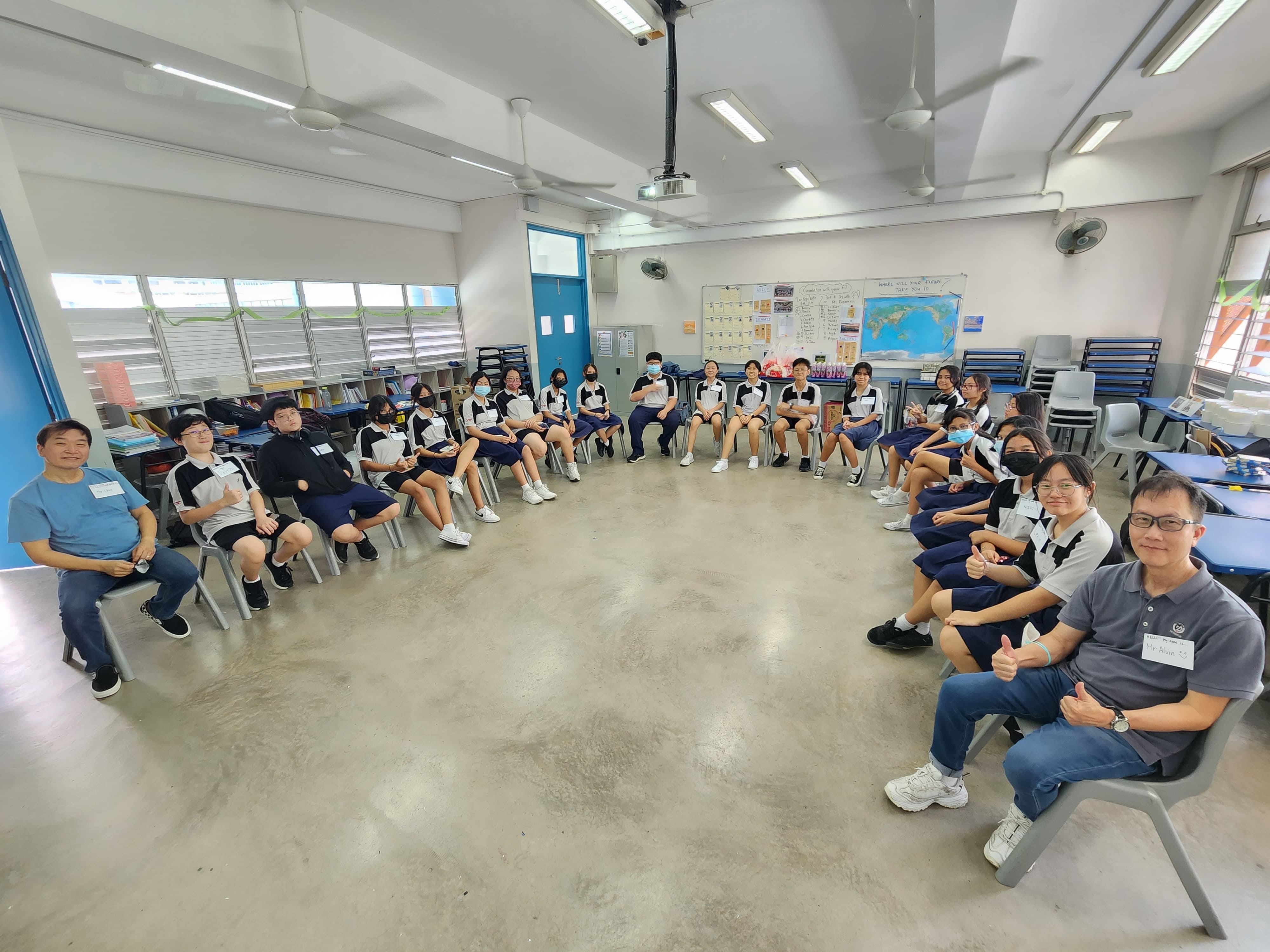 Students in navy and white uniforms sit in a large circle with two adults in a bright classroom.
